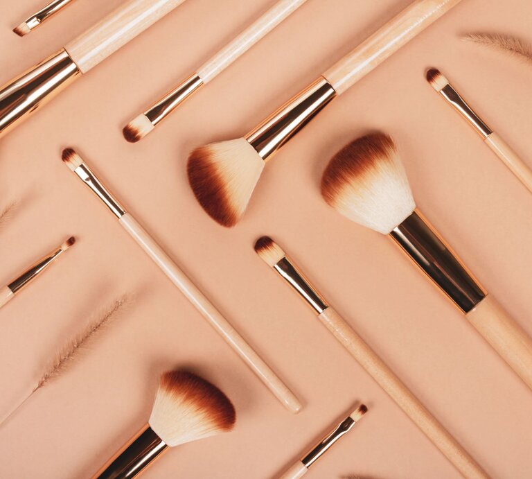 Verschiedene Make-up-Pinsel mit hellen Holzgriffen und braunen Borsten liegen ordentlich auf einem beigen Untergrund. | © GettyImages/Iana Kunitsa