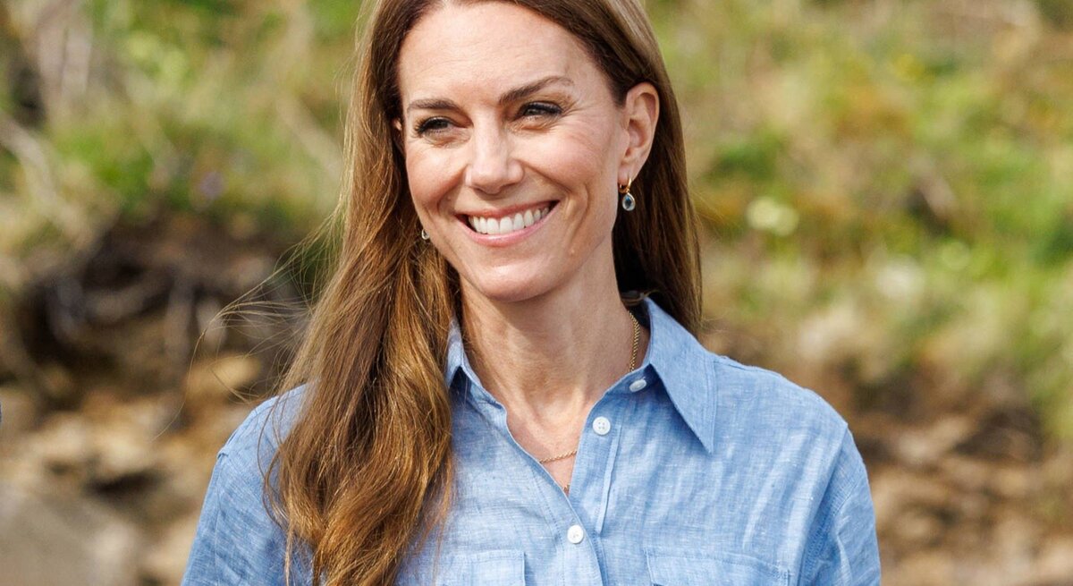Lachende Prinzessin Kate mit langen braunen Haaren in hellblauer Bluse steht im Grünen vor verschwommenem Waldboden.