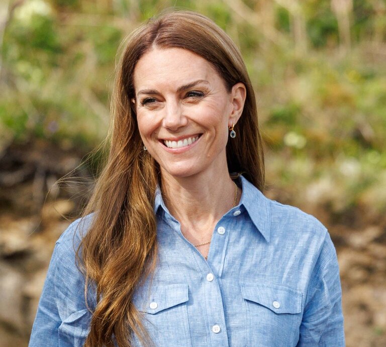 Lachende Prinzessin Kate mit langen braunen Haaren in hellblauer Bluse steht im Grünen vor verschwommenem Waldboden. | © IMAGO / Spotlight Royal