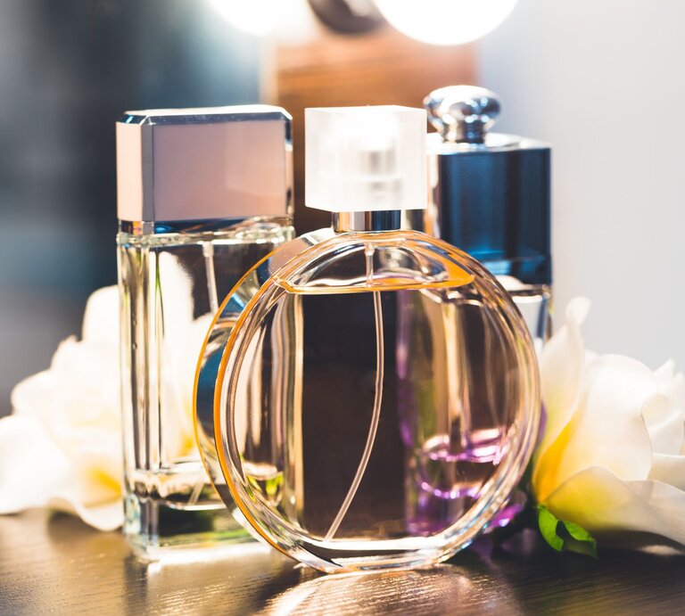 Drei elegante Parfümflakons in verschiedenen Formen stehen auf einem dunklen Tisch, umgeben von weißen Blumen und warmem Licht. | © Adobe Stock/ fotofabrika