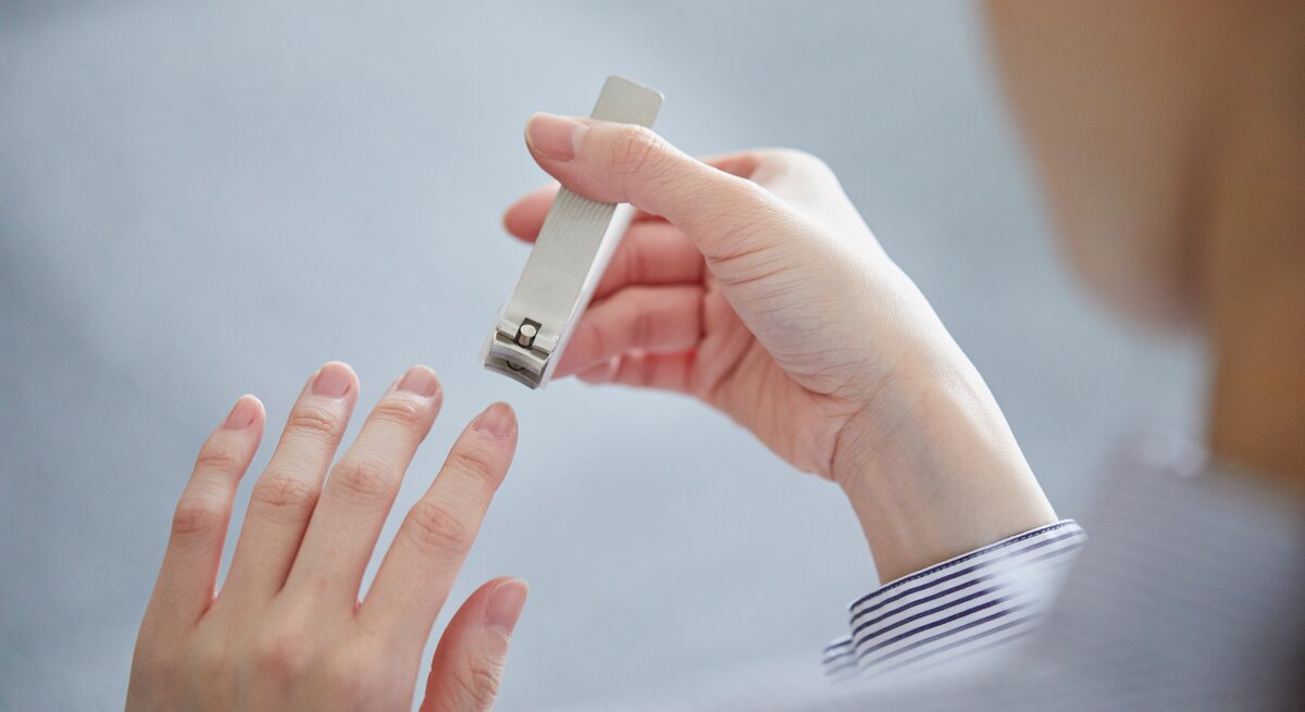 Eine Person mit gestreifter Manschette hält einen silbernen Nagelknipser und schneidet Fingernägel vor neutral grauem Hintergrund.