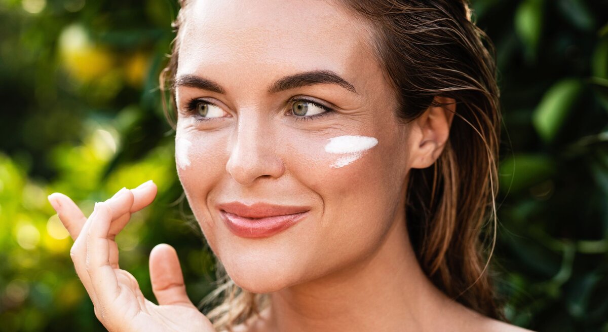 Lächelnde junge Frau mit feuchtem Haar trägt Sonnencreme auf ihre Wange auf, grüne Pflanzen im Hintergrund.