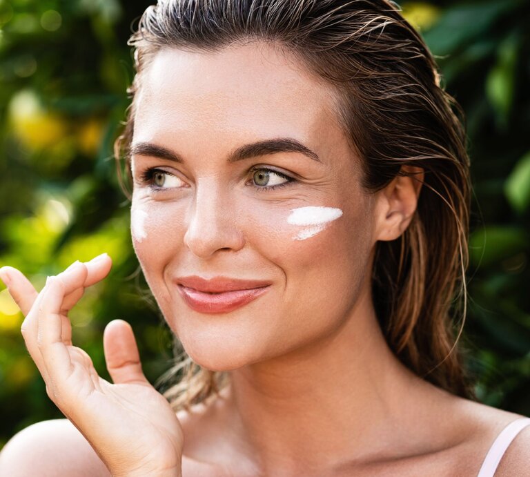 Lächelnde junge Frau mit feuchtem Haar trägt Sonnencreme auf ihre Wange auf, grüne Pflanzen im Hintergrund. | © Getty Images/ Jun