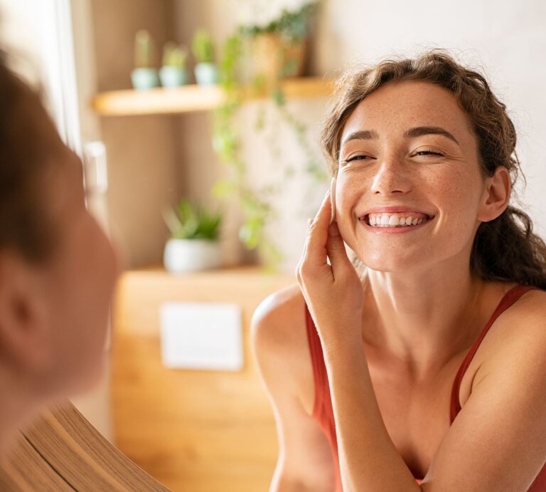 Das Bild zeigt eine junge Frau mit einem herzlichen und offenen Lächeln, die einem anderen Menschen gegenüber sitzt. Die Frau trägt scheinbar keine schwere Make-up-Schicht, aber man kann deutliche, natürlich aussehende Sommersprossen auf ihren Wangen und ihrer Nase erkennen. Diese könnten echte Sommersprossen sein oder sorgfältig mit Make-up geschaffen worden sein, um einen spielerischen und sommerlichen Look zu erzeugen, was darauf hindeutet, dass es sich um den Trend des Schminkens von Fake Sommersprossen handeln könnte. Die Frau hat die Haare locker hochgesteckt und trägt ein einfaches, rotes Tanktop, was den Eindruck eines entspannten und gemütlichen Umfelds verstärkt. Das Umfeld zeigt ein warmes, wohnliches Interieur mit unscharfem Hintergrund, der auf eine gut beleuchtete Küche oder ein Wohnzimmer hindeutet. | © gettyimages/	Ridofranz