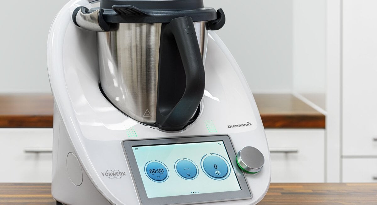 Das Bild zeigt einen modernen Thermomix Küchenhelfer von Vorwerk auf einer Küchenarbeitsplatte. Das Gerät verfügt über ein integriertes digitales Bedienfeld mit einem Touchscreen, auf dem eine Uhr und Bedienelemente zu sehen sind. Der Thermomix besteht aus einer Basisstation mit einem Display und Bedienelementen sowie einem großen Mixtopf aus Edelstahl mit einem schwarzen Griff und einem Messer im Inneren. Auf der rechten Seite des Geräts ist eine Öffnung für den Varoma-Aufsatz oder andere Zubehörteile zu erkennen. Das Design ist schlicht und in einem sauberen Weiß gehalten, mit dem Vorwerk-Logo deutlich sichtbar an der Vorderseite. Der Hintergrund ist schlicht und lenkt nicht von dem Küchengerät ab, was den Fokus auf den Thermomix richtet.