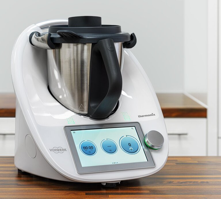 Das Bild zeigt einen modernen Thermomix Küchenhelfer von Vorwerk auf einer Küchenarbeitsplatte. Das Gerät verfügt über ein integriertes digitales Bedienfeld mit einem Touchscreen, auf dem eine Uhr und Bedienelemente zu sehen sind. Der Thermomix besteht aus einer Basisstation mit einem Display und Bedienelementen sowie einem großen Mixtopf aus Edelstahl mit einem schwarzen Griff und einem Messer im Inneren. Auf der rechten Seite des Geräts ist eine Öffnung für den Varoma-Aufsatz oder andere Zubehörteile zu erkennen. Das Design ist schlicht und in einem sauberen Weiß gehalten, mit dem Vorwerk-Logo deutlich sichtbar an der Vorderseite. Der Hintergrund ist schlicht und lenkt nicht von dem Küchengerät ab, was den Fokus auf den Thermomix richtet. | © Adobe Stock/ Mathias Weil