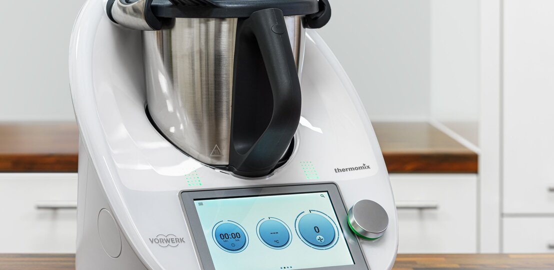 Das Bild zeigt einen modernen Thermomix Küchenhelfer von Vorwerk auf einer Küchenarbeitsplatte. Das Gerät verfügt über ein integriertes digitales Bedienfeld mit einem Touchscreen, auf dem eine Uhr und Bedienelemente zu sehen sind. Der Thermomix besteht aus einer Basisstation mit einem Display und Bedienelementen sowie einem großen Mixtopf aus Edelstahl mit einem schwarzen Griff und einem Messer im Inneren. Auf der rechten Seite des Geräts ist eine Öffnung für den Varoma-Aufsatz oder andere Zubehörteile zu erkennen. Das Design ist schlicht und in einem sauberen Weiß gehalten, mit dem Vorwerk-Logo deutlich sichtbar an der Vorderseite. Der Hintergrund ist schlicht und lenkt nicht von dem Küchengerät ab, was den Fokus auf den Thermomix richtet. | © Adobe Stock/ Mathias Weil