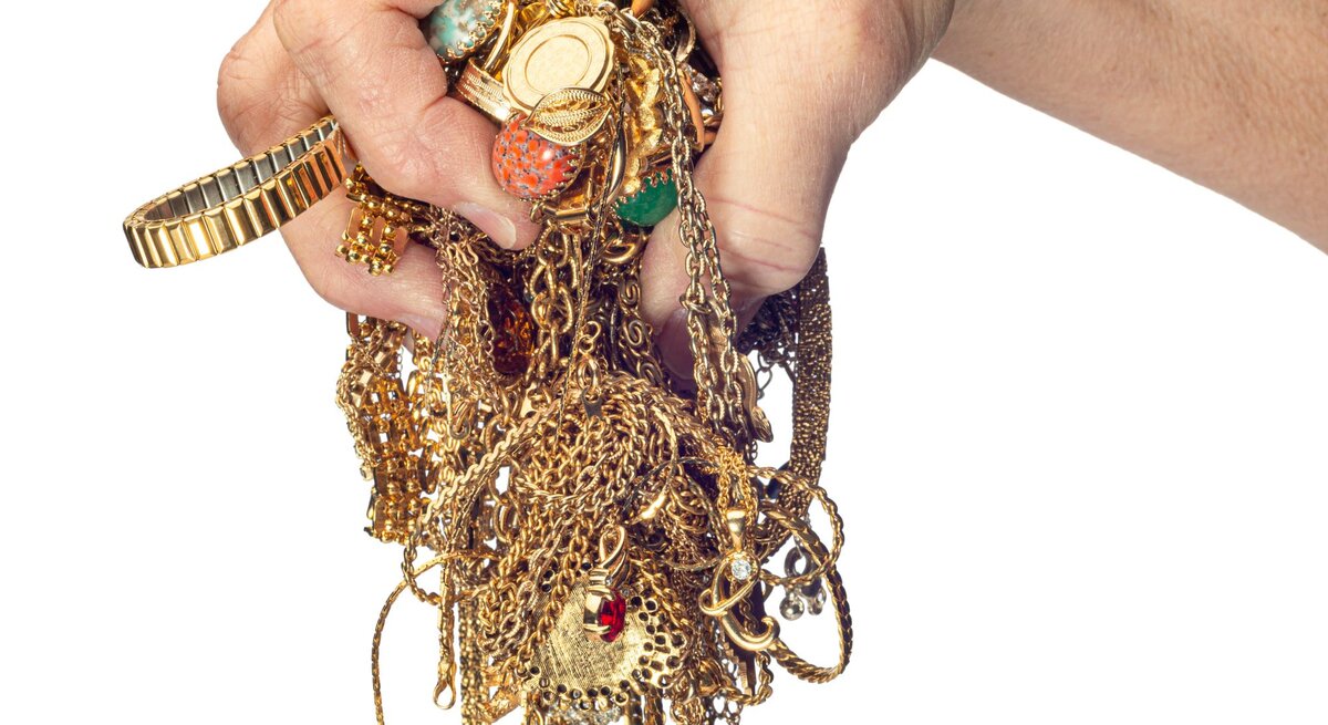 Hand, die einen Haufen von verknoteten Goldketten, Armbändern und anderen Schmuckstücken hält. Die Schmuckstücke sind stark ineinander verwickelt.