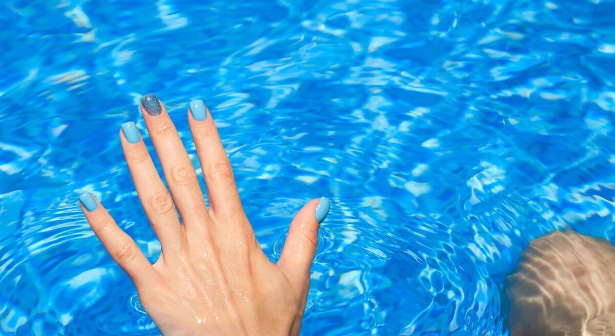 Hand mit blau lackierten Nägeln und glitzerndem Akzentfinger taucht in klares, funkelndes Poolwasser bei sonnigem Licht.