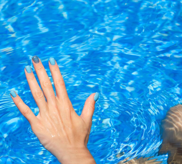Hand mit blau lackierten Nägeln und glitzerndem Akzentfinger taucht in klares, funkelndes Poolwasser bei sonnigem Licht. | © Adobe Stock/ Philipp Berezhnoy