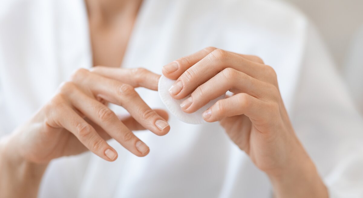 Hände einer Person, die eine weiße Wattepadscheibe zwischen Daumen und Zeigefinger hält, unscharfer Oberkörper in weißem Hemd im Hintergrund.