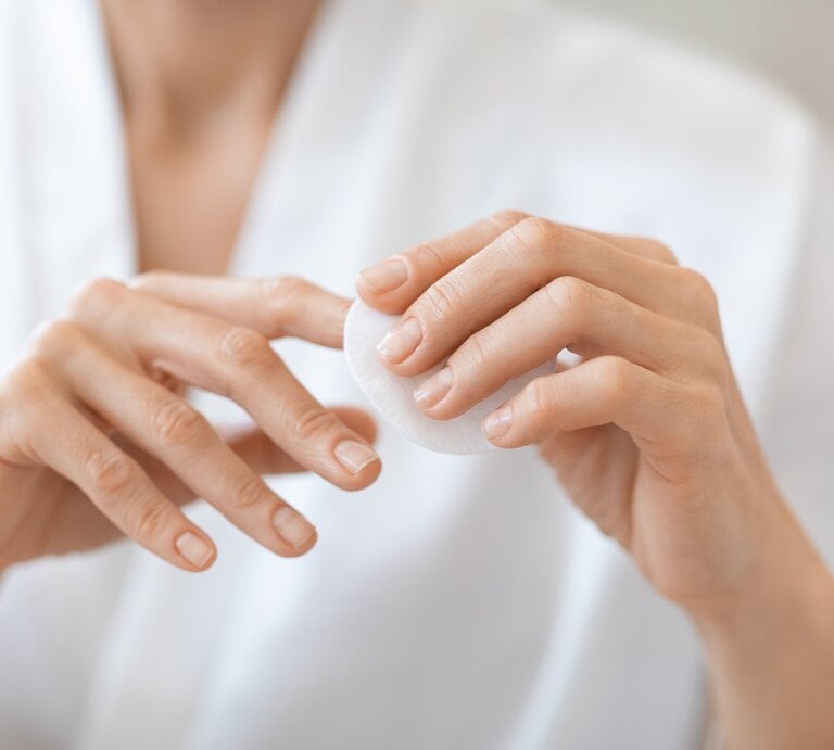 Hände einer Person, die eine weiße Wattepadscheibe zwischen Daumen und Zeigefinger hält, unscharfer Oberkörper in weißem Hemd im Hintergrund. | © Adobe Stock/ Prostock-studio