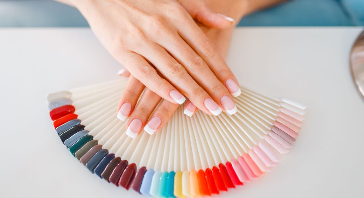 Nahaufnahme von Händen mit dezentem French Maniküre-Look, die einen Nagellack-Farbfächer präsentieren.