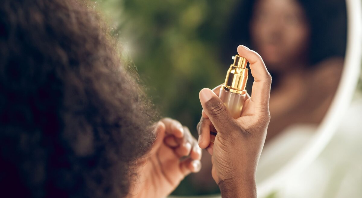 Frau trägt vor einem Spiegel Parfüm auf. Sie hält den Parfümflakon elegant in der Hand und richtet ihn auf ihren Hals. Ihr Gesicht ist im Hintergrund im Spiegel verschwommen sichtbar, was einen privaten und alltäglichen Moment einfängt.