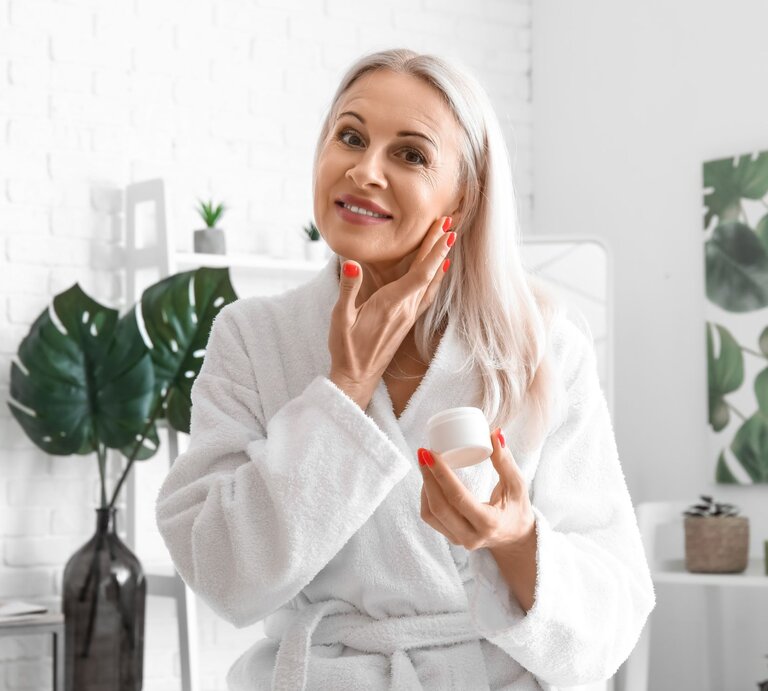 Ältere Frau mit langem grauem Haar cremt lächelnd ihr Gesicht ein, trägt weißen Bademantel in heller Wohnung. | © Adobe Stock/ Pixel-Shot