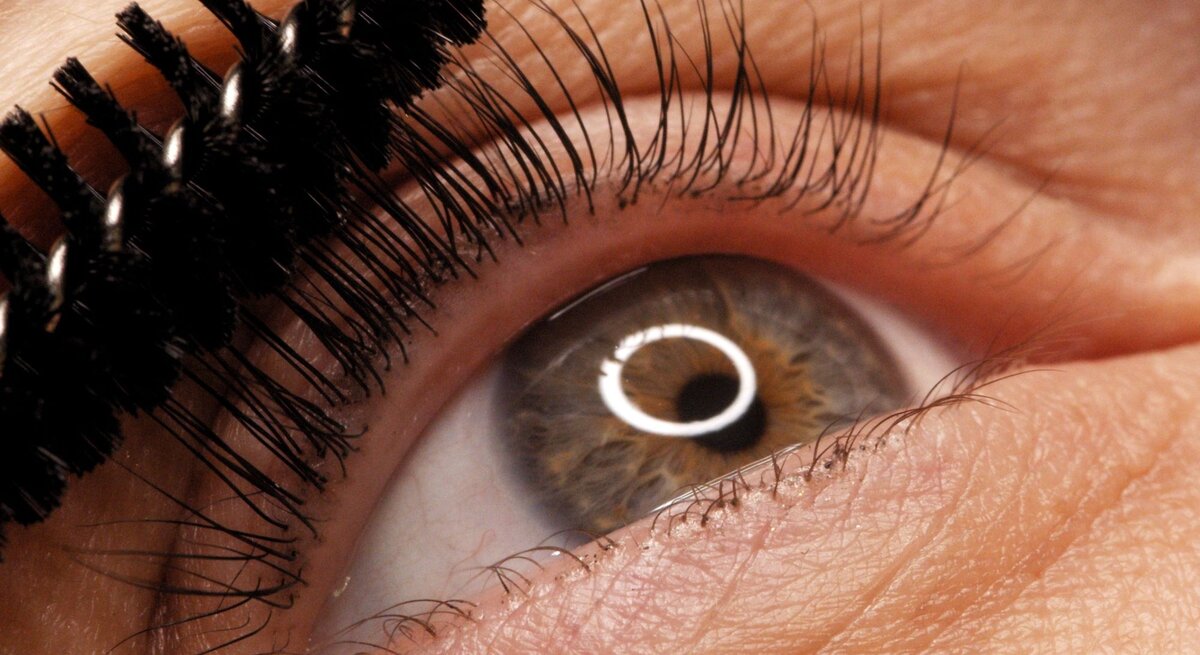 Nahaufnahme eines Auges mit brauner Iris, das mit einer Mascara-Bürste die oberen Wimpern tuscht.
