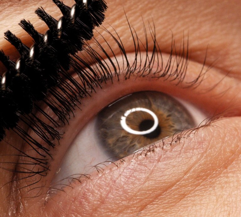 Nahaufnahme eines Auges mit brauner Iris, das mit einer Mascara-Bürste die oberen Wimpern tuscht. | © Adobe Stock/ Valua Vitaly