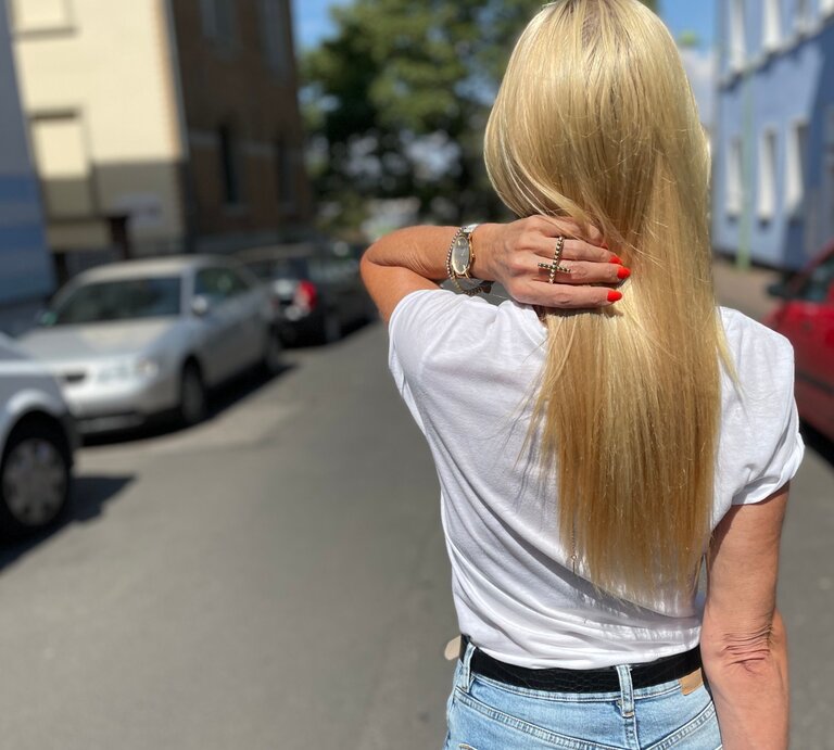 Eine Frau mit glattem, langen, blonden Haar, rotem Nagellack und Jeans streicht sich das Haar in einer ruhigen Wohnstraße zurück. | © Renate Zott
