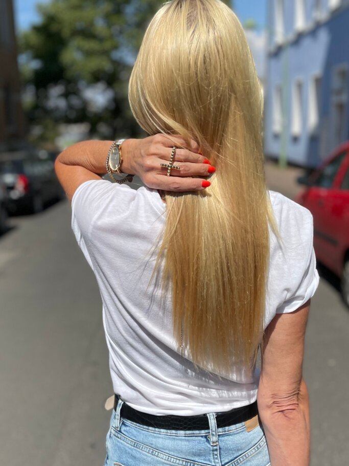 Eine Frau mit glattem, langen, blonden Haar, rotem Nagellack und Jeans streicht sich das Haar in einer ruhigen Wohnstraße zurück. | © Renate Zott
