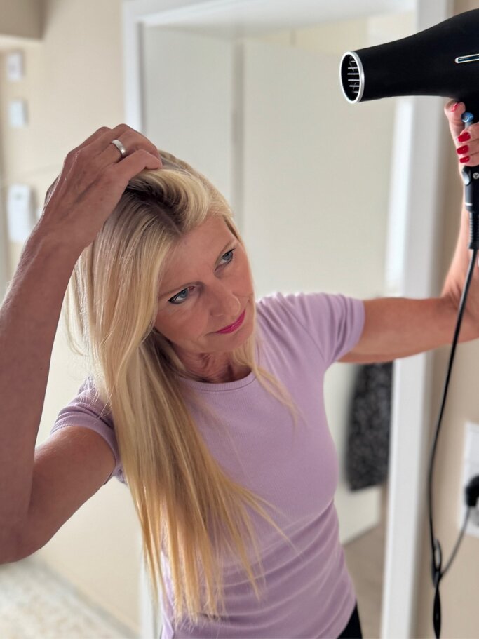 Blonde Frau im engen lila Oberteil verwendet einen schwarzen Föhn, während sie mit der Hand ihr Haar anhebt, in einem hellen Raum. | © Renate Zott