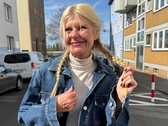 Renate Zott mit geflochtenen Zöpfen, blauer Jeansjacke und hellen Lippen lächelt sanft bei Sonnenschein zwischen Wohnhäusern. | © Renate Zott