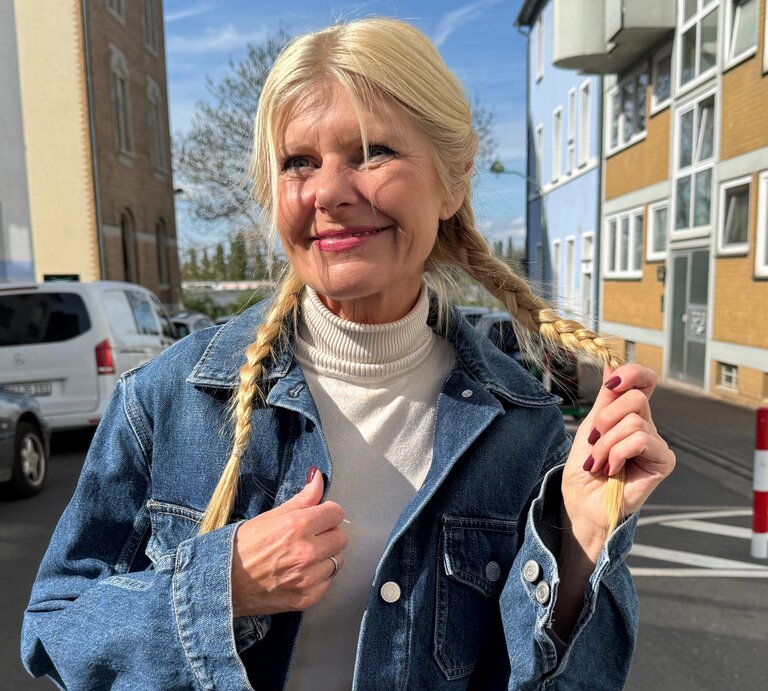 Renate Zott mit geflochtenen Zöpfen, blauer Jeansjacke und hellen Lippen lächelt sanft bei Sonnenschein zwischen Wohnhäusern. | © Renate Zott