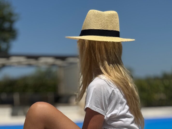 Renate Zott mit Strohhut und weißem T-Shirt sitzt am Pool und blickt in einen sonnigen Garten mit blauem Himmel. | © Renate Zott 