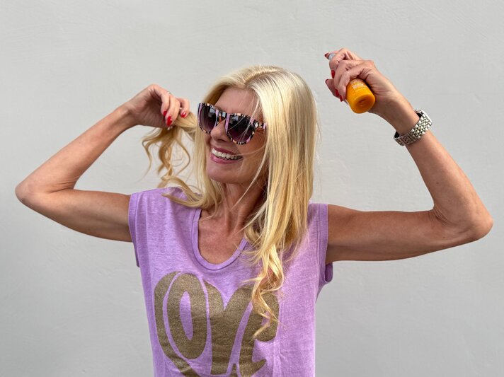 Renate Zott mit langen blonden Haaren sprüht sich mit Sonnenspray ein, trägt lila Top und bunte Sonnenbrille. | © Renate Zott