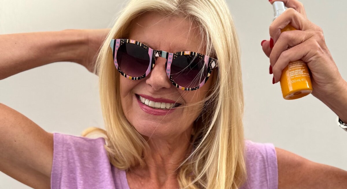 Renate Zott mit offenem blondem Haar posiert in pinker Sonnenbrille und lila Shirt, hält Sprayflasche hoch.
