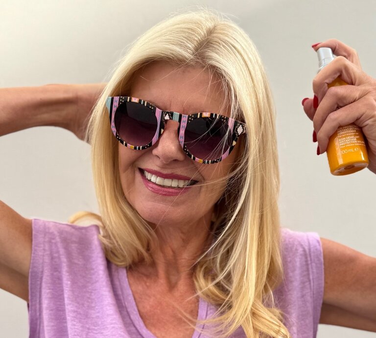 Renate Zott mit offenem blondem Haar posiert in pinker Sonnenbrille und lila Shirt, hält Sprayflasche hoch. | © Renate Zott