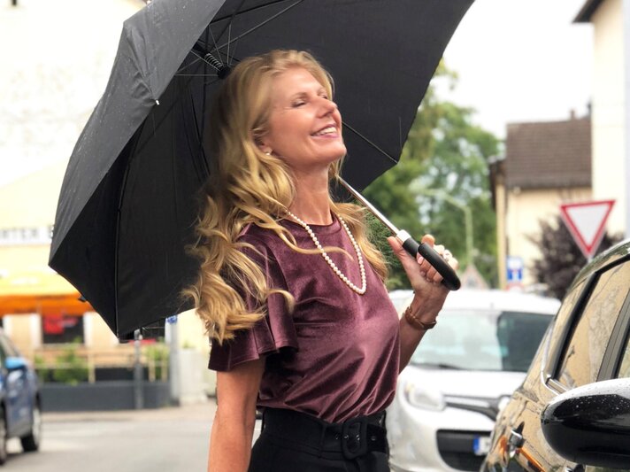 Renate Zott mit Perlenkette und violetter Bluse hält fröhlich einen schwarzen Schirm in regennasser Straße. | © Renate Zott