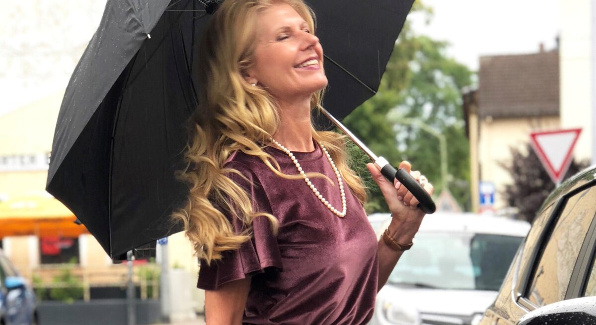 Renate Zott mit Perlenkette und violetter Bluse hält fröhlich einen schwarzen Schirm in regennasser Straße.