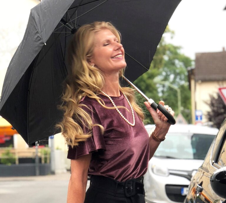 Renate Zott mit Perlenkette und violetter Bluse hält fröhlich einen schwarzen Schirm in regennasser Straße. | © Renate Zott