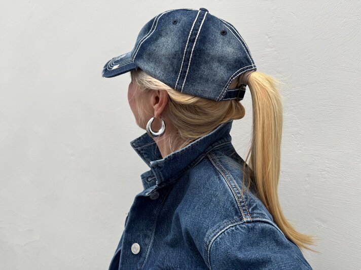 Blondhaarige Renate Zott mit Pferdeschwanz und silbernen Creolen trägt Jeansjacke und Baseballcap, im Profil vor grauer Wand. | © Renate Zott