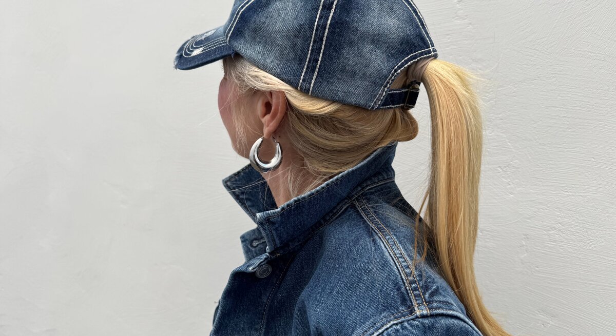 Blondhaarige Renate Zott mit Pferdeschwanz und silbernen Creolen trägt Jeansjacke und Baseballcap, im Profil vor grauer Wand.