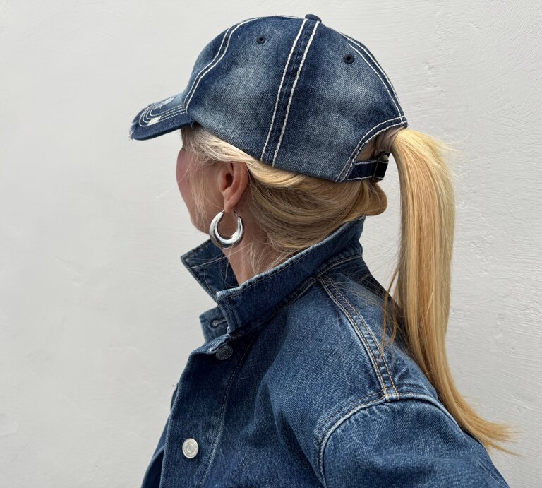 Blondhaarige Renate Zott mit Pferdeschwanz und silbernen Creolen trägt Jeansjacke und Baseballcap, im Profil vor grauer Wand. | © Renate Zott