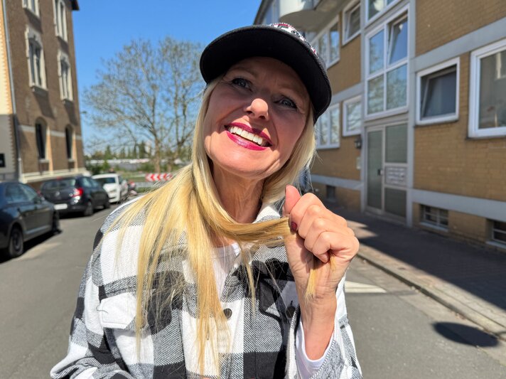 Lachende Renate Zott mit Cap, pinkem Lippenstift und kariertem Mantel steht auf sonniger Straße zwischen Alt- und Neubauten. | © Renate Zott