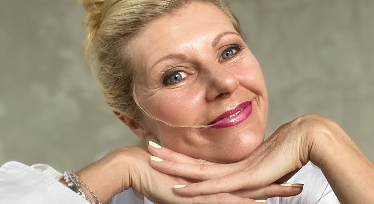 Blondhaarige ältere Frau (Renate Zott) mit Dutt lächelt sanft, stützt das Kinn auf die Hände und trägt rosa Lippenstift sowie weiße Bluse.