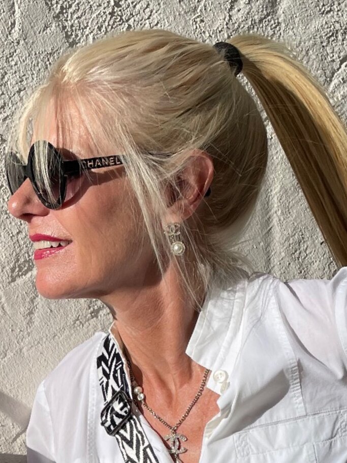 Blondhaarige Renate Zott mit hohem Pferdeschwanz und Chanel-Sonnenbrille blickt lächelnd in die Sonne vor rauer weißer Wand. | © Renate Zott