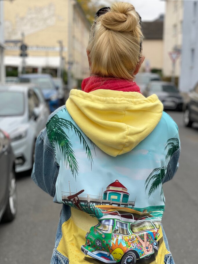 Rückansicht von Renate Zott mit klassischem Dutt, farbenfrohem Kapuzenpulli und Jeansjacke im urbanen Umfeld bei bedecktem Himmel. | © Renate Zott