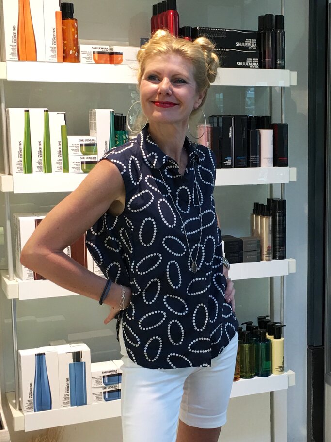 Blondhaarige Renate Zott mit Doppeldutt und rotem Lippenstift posiert fröhlich vor Regal mit Pflegeprodukten in eleganter Sommerkleidung. | © Renate Zott