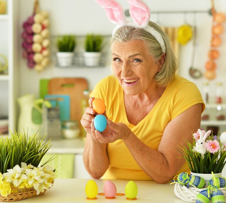 Eine ältere Frau mit grauen Haaren und Hasenohren-Stirnband hält bunt gefärbte Ostereier und lächelt fröhlich in einer frühlingshaft dekorierten Küche. | © Shutterstock/Ruslan Huzau