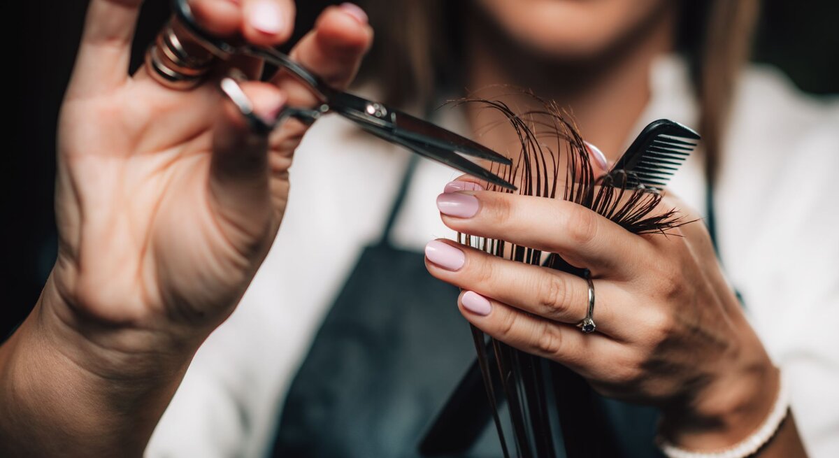 Nahaufnahme von Händen einer Friseurin mit Schere und Kamm beim präzisen Schneiden brauner Haare. Trendfrisuren 2025