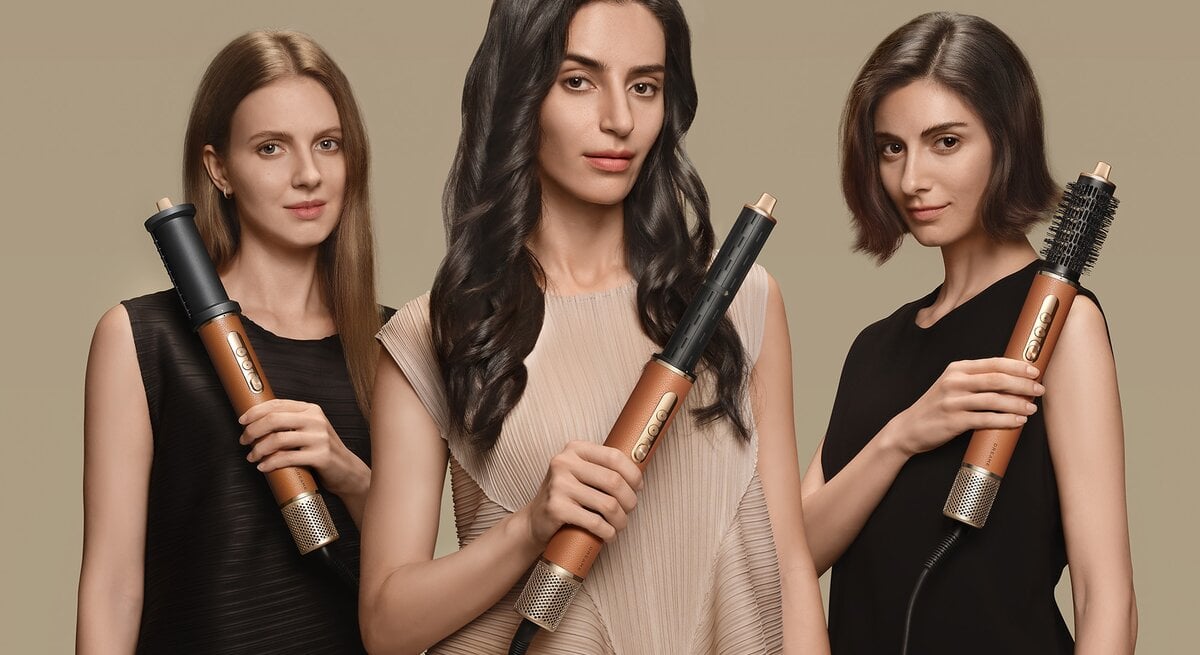 Drei junge Frauen mit glatten, welligen und kurzen Haaren präsentieren in eleganten Kleidern goldene Haarstyling-Geräte vor beigem Hintergrund