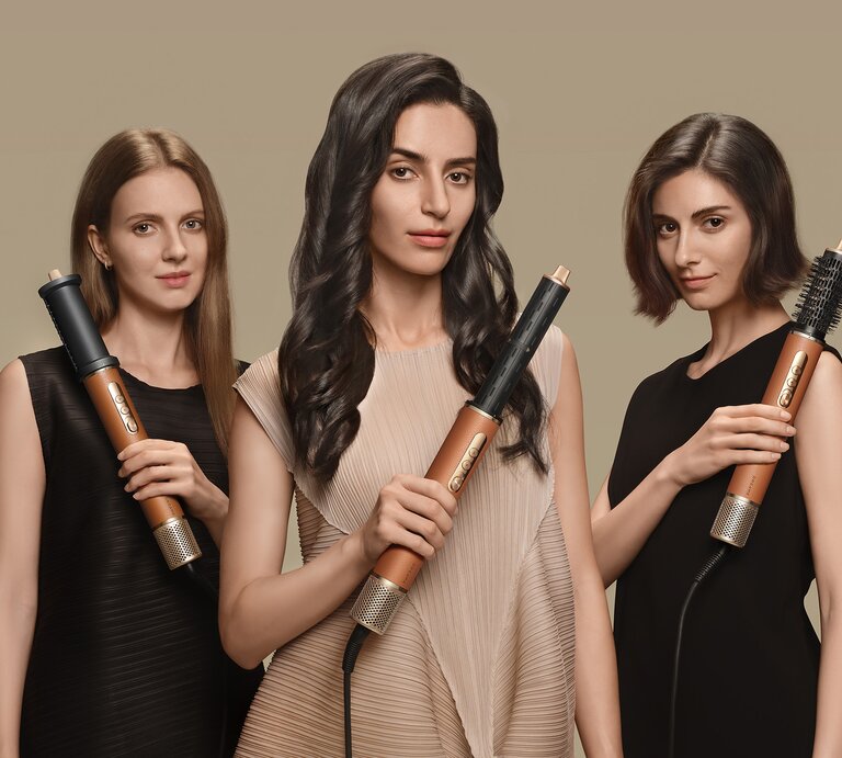 Drei junge Frauen mit glatten, welligen und kurzen Haaren präsentieren in eleganten Kleidern goldene Haarstyling-Geräte vor beigem Hintergrund | © Dreame