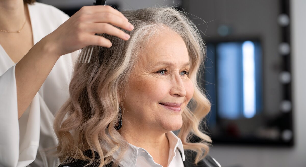 Eine Hand im weißen Ärmel kämmt weiche, blonde Locken; im Hintergrund ein dunkel eingerichteter Friseursalon.