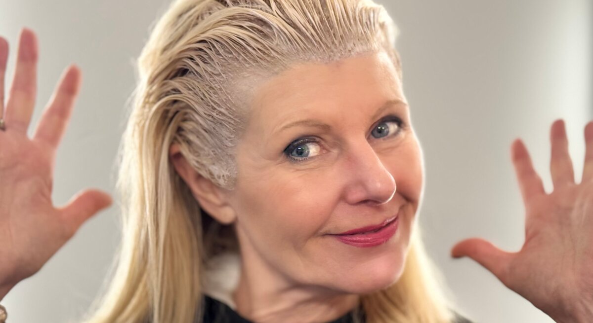 Renate Zott beim graue Haare färben, lächelt in die Kamera und hebt fröhlich die Hände.