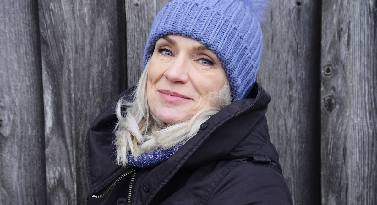 Eine ältere Frau mit blondem Haar, blauer Strickmütze und schwarzer Jacke lächelt sanft vor einer verwitterten Holzwand.
