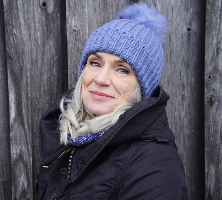 Eine ältere Frau mit blondem Haar, blauer Strickmütze und schwarzer Jacke lächelt sanft vor einer verwitterten Holzwand. | © Getty Images/ Daniel Attia