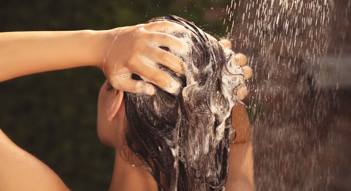 Nahaufnahme einer Person mit nassen Haaren, die sich unter fließendem Wasser die Haare mit Shampoo einschäumt und die Sandich-Methode verwendet.
